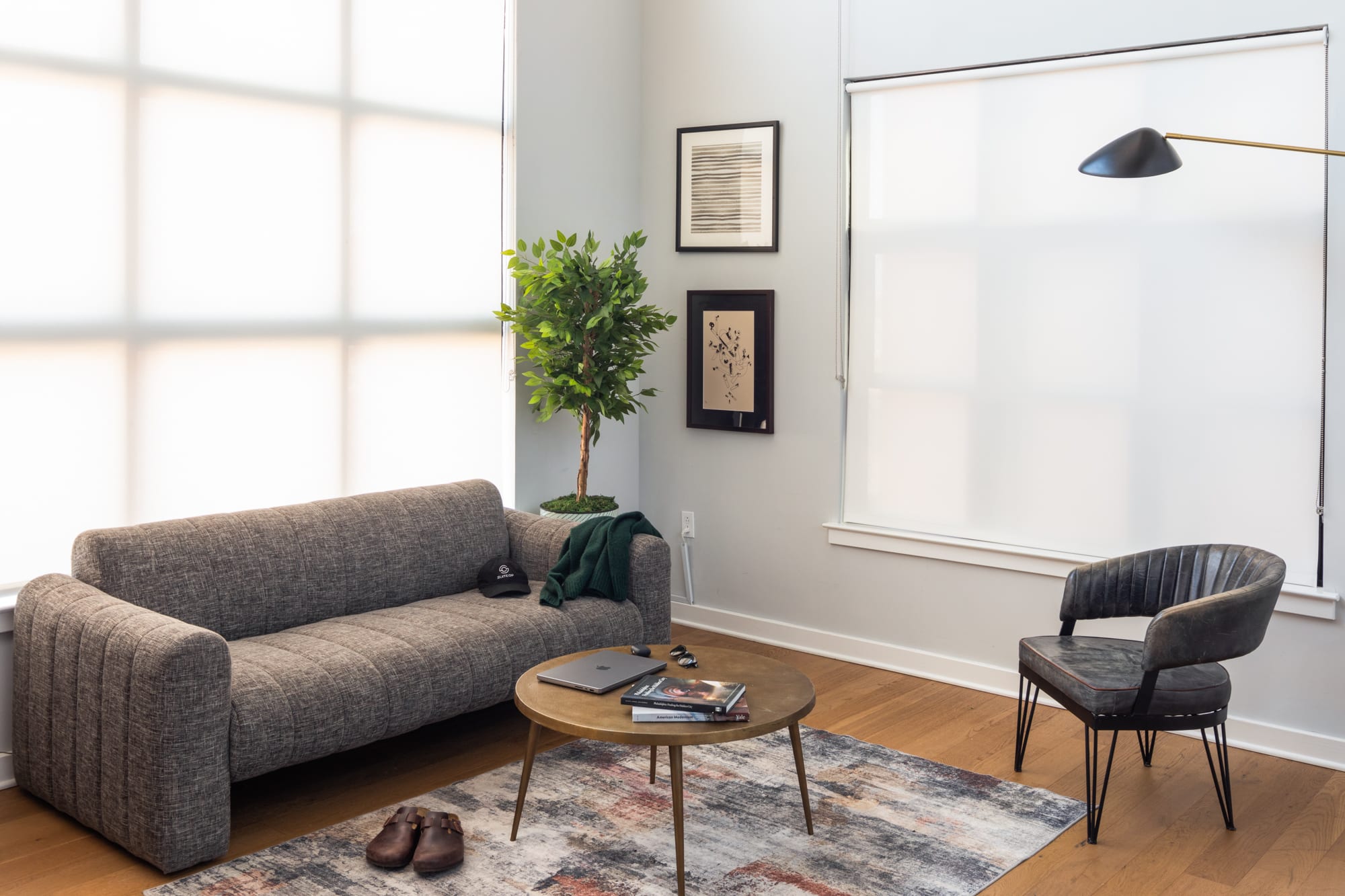 A modern living room with light hardwood floors, a textured gray sofa, a round gold coffee table on a patterned rug, a dark leather armchair, and a potted plant by large windows with white roller shades.