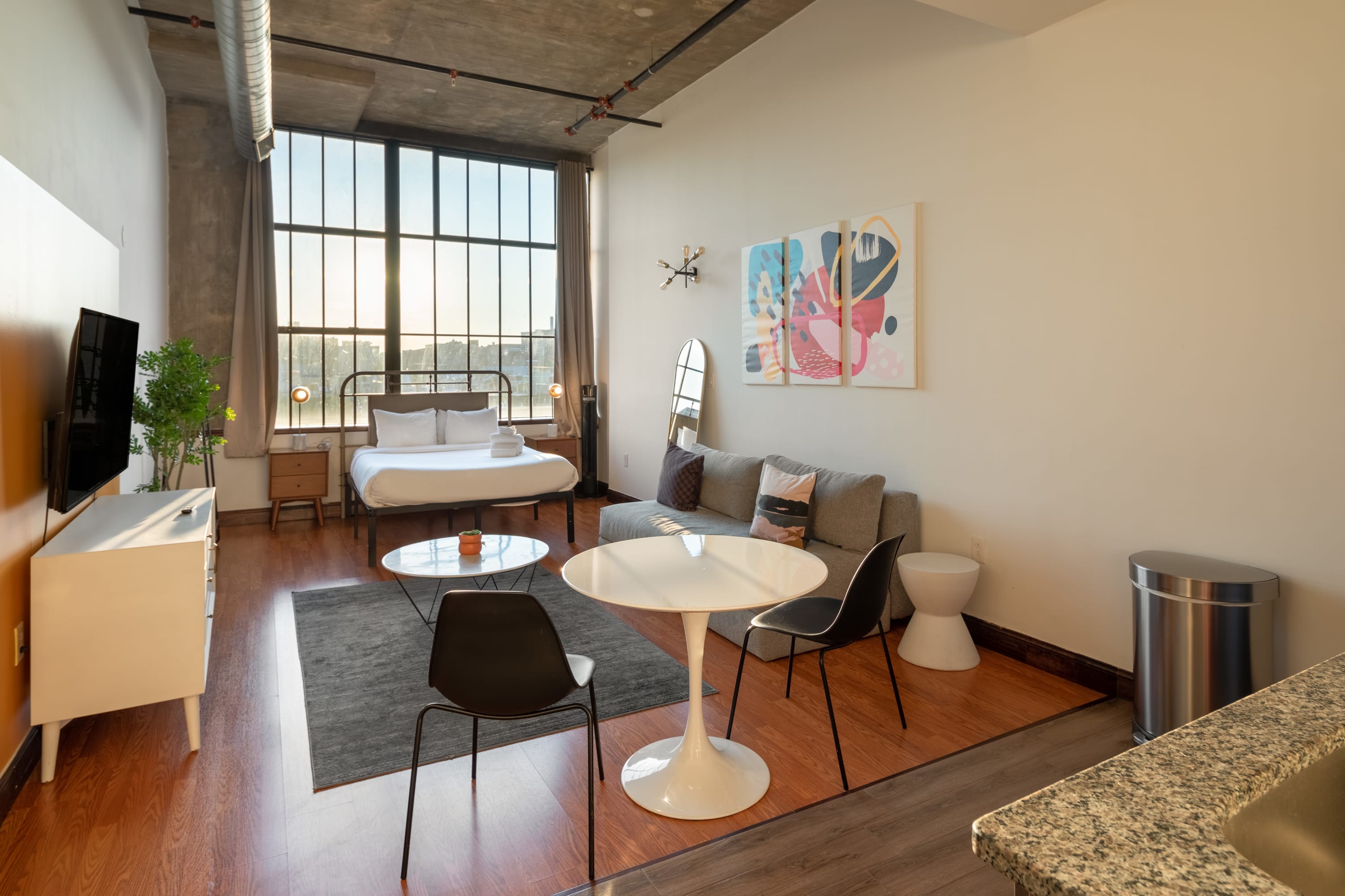 An open-concept studio apartment with a queen bed near large industrial windows, a gray sofa, a white round dining table with black chairs, and a compact kitchen visible in the background, all on warm hardwood floors.