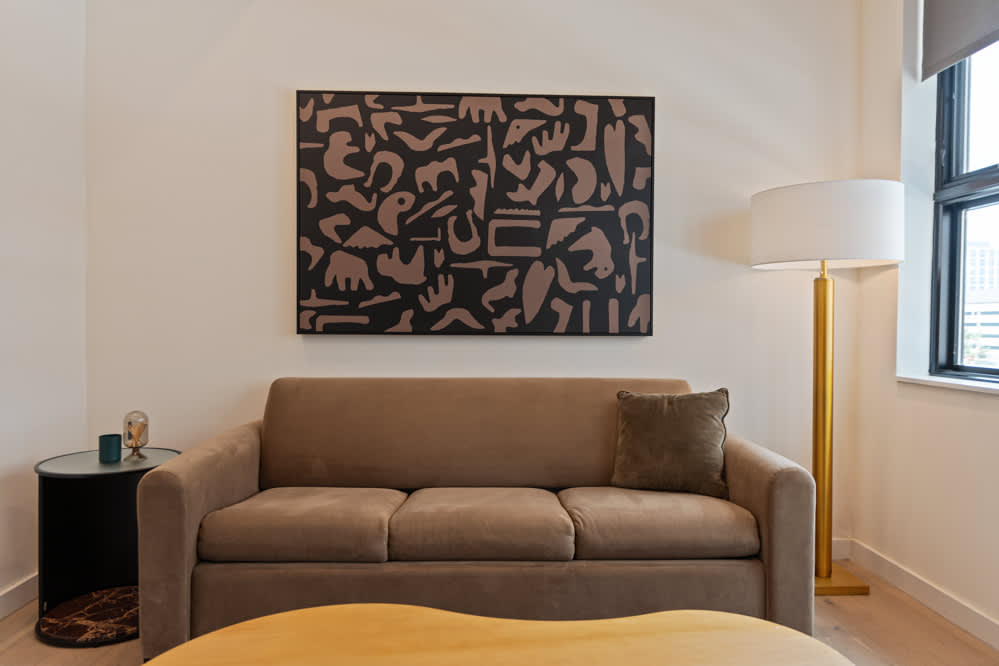 A contemporary living room features a comfortable light brown sofa with a dark throw pillow, a unique kidney-shaped wooden coffee table, and a large abstract black and brown artwork on the wall. A tall gold floor lamp stands beside the sofa.
