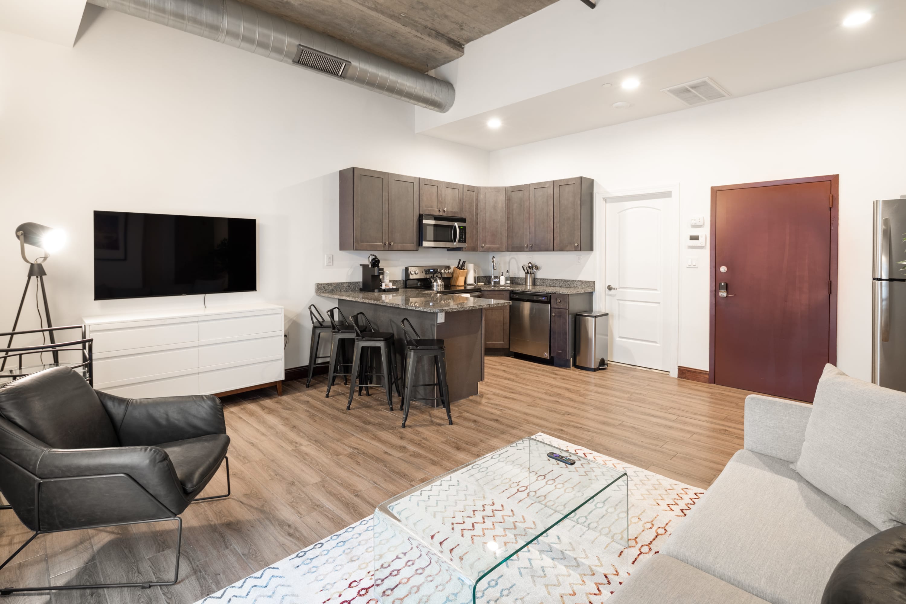A spacious open-concept living room and kitchen with light wood flooring and a concrete ceiling. The living area includes a grey sofa, a black leather armchair, and a wall-mounted TV, while the adjacent kitchen has dark cabinets, granite countertops, and a breakfast bar with stools.