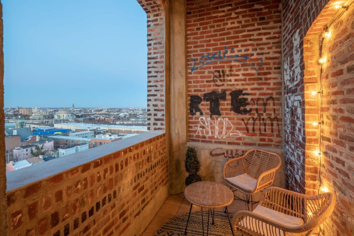 Cozy balcony featuring exposed brick walls with graffiti, string lights, and two wicker chairs with a small round table, overlooking a sprawling city landscape.