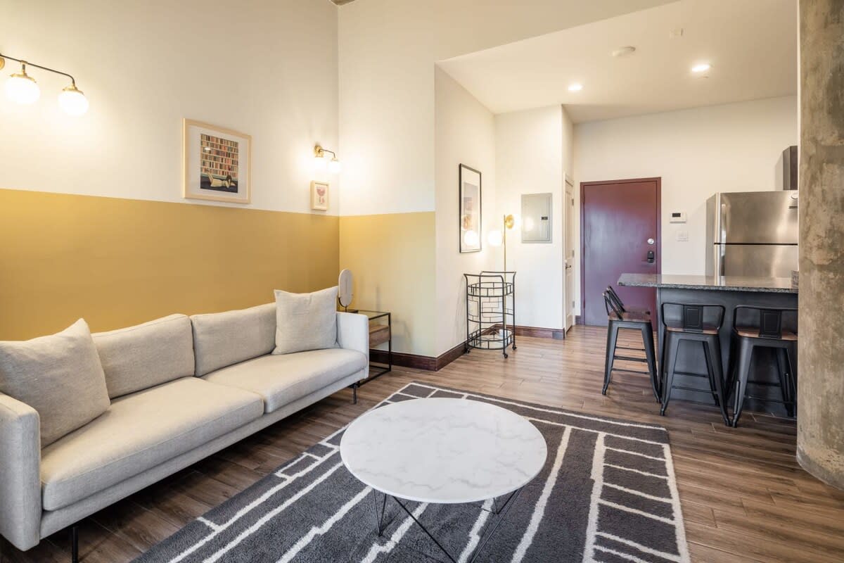 An open-concept living area with a light gray sofa, a round marble-top coffee table, and a geometric rug. A yellow and white accent wall adds a pop of color, and a kitchen bar with stools is visible in the background.