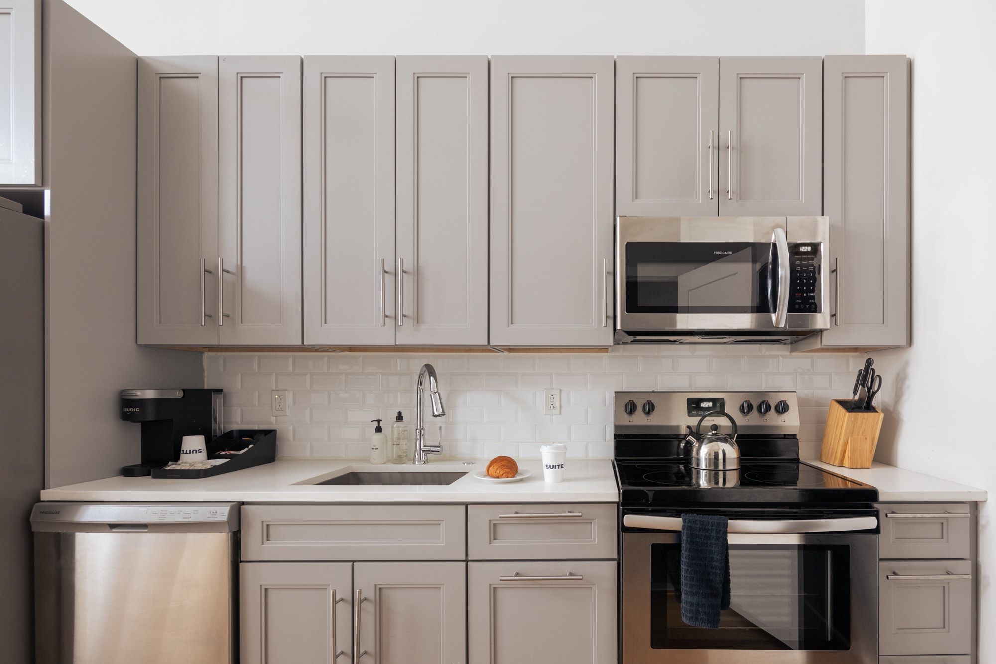 A modern kitchen featuring light gray shaker-style cabinets, white subway tile backsplash, and stainless steel appliances including a microwave, dishwasher, and range. A white countertop holds a sink, coffee maker, and kettle.
