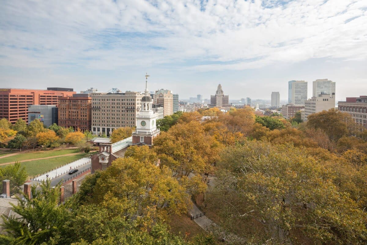 Panoramic aerial view of a historic city park with vibrant autumn trees, surrounded by classic and modern city buildings under a partly cloudy sky.