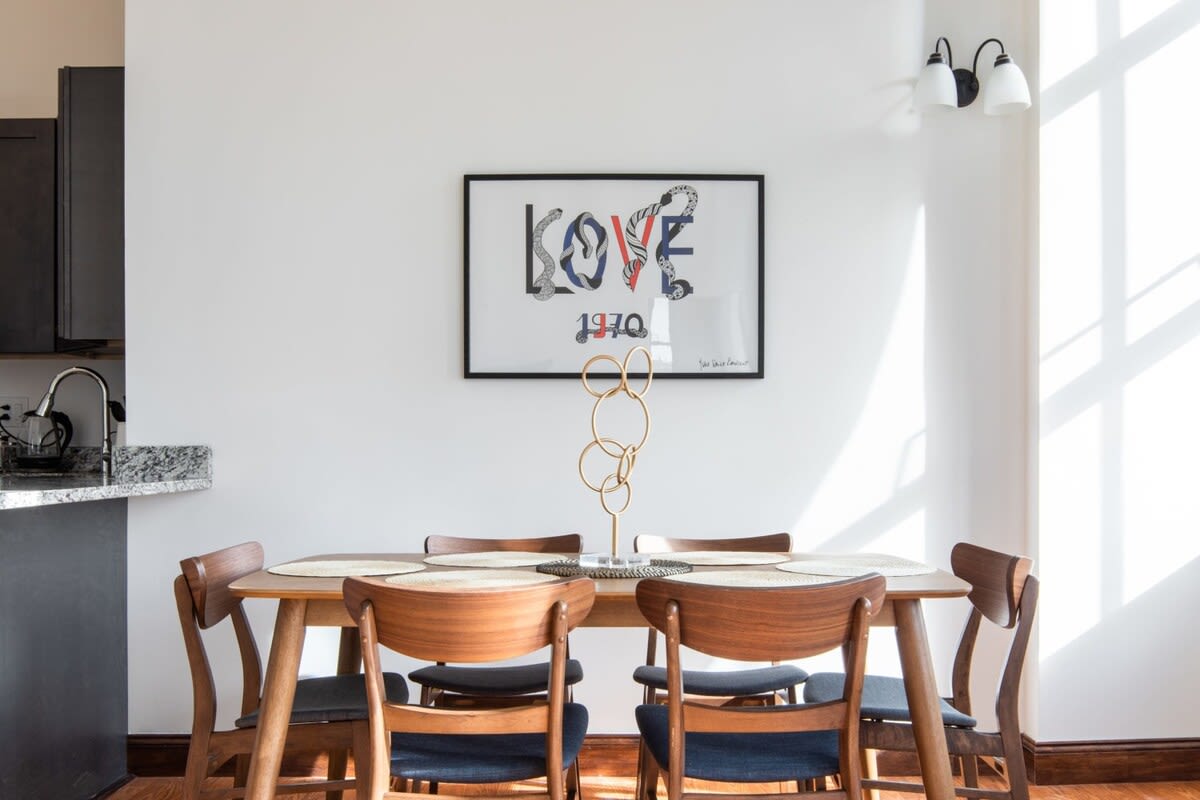 A stylish dining area centered around a light wood table with six mid-century modern chairs upholstered in dark blue. A framed 'LOVE 1970' artwork adorns the white wall, with a unique gold ring sculpture as a table centerpiece.