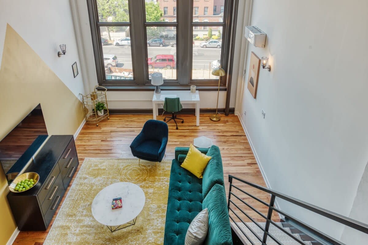 Overhead view of a bright loft living room with large industrial-style windows, a teal sofa, and a white desk with a green chair. The space features hardwood floors and a yellow patterned area rug.
