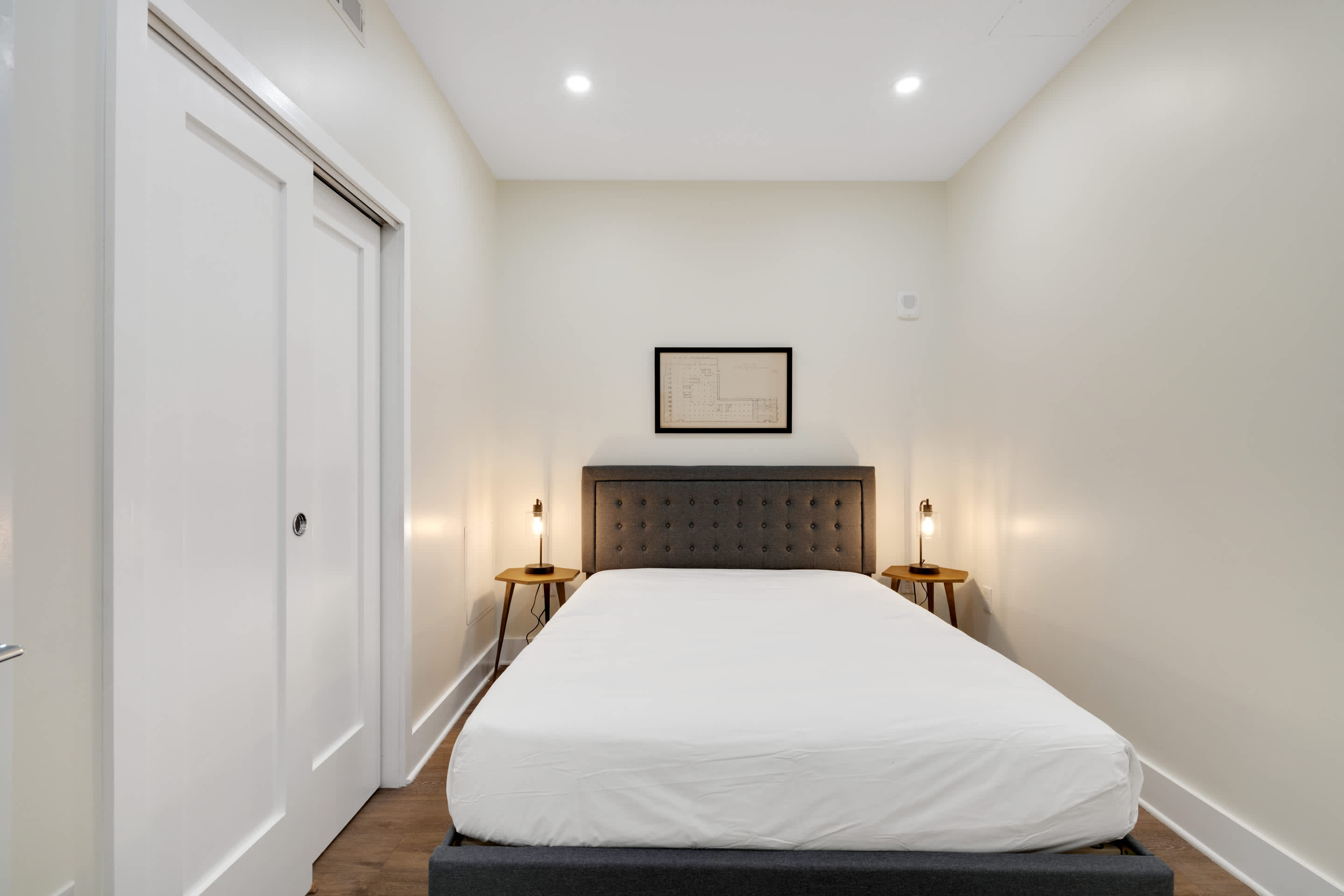 Cozy bedroom featuring a dark gray upholstered bed with a tufted headboard, flanked by two small round wooden nightstands with modern lamps. A framed blueprint-style artwork hangs above the bed, and white sliding closet doors are visible on the left.