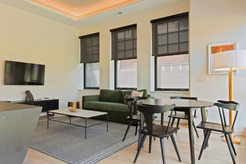 Bright living area with a dark green sofa, a white marble-top coffee table, a striped rug, and a round black dining table with matching chairs, all illuminated by natural light from three large windows with dark roller shades.