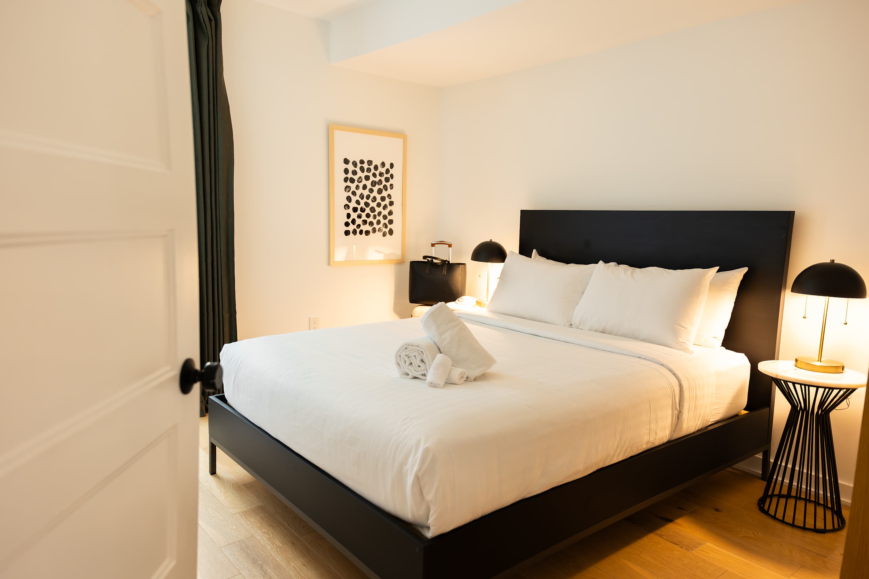 A cozy bedroom with a black platform bed and white linens, flanked by modern nightstands with lamps, and a framed abstract art piece above one nightstand, set on light hardwood floors.