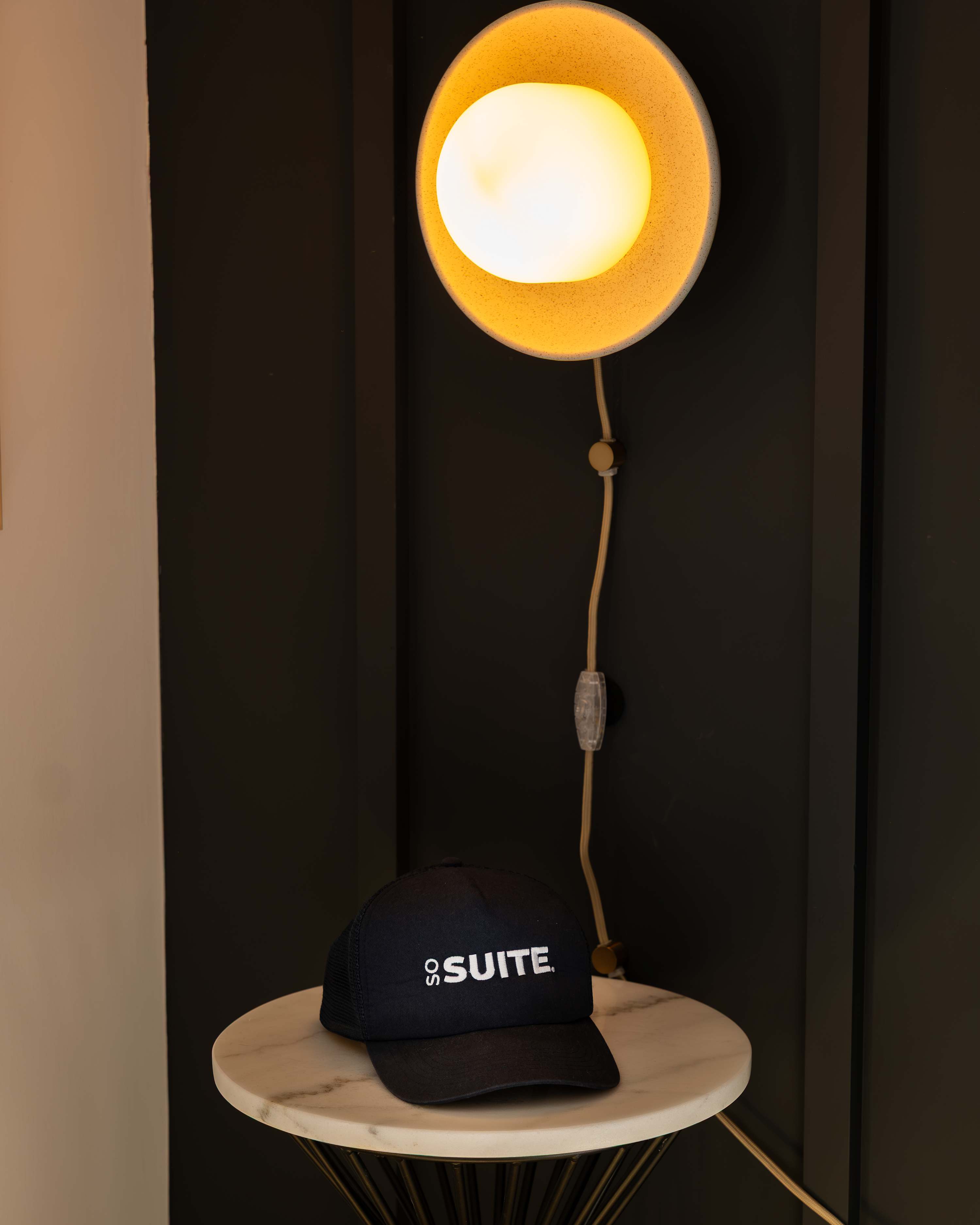 A close-up of a marble-top nightstand featuring a modern wall-mounted lamp with a circular glow and a black "SoSuite" baseball cap resting on the surface. The background shows a dark paneled wall.