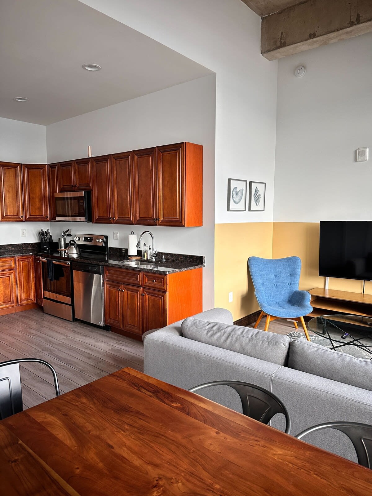 Modern kitchen with wooden cabinetry, granite countertops, and stainless steel appliances, seamlessly flowing into a living area with a light gray sofa and a blue accent chair, under a high ceiling with exposed ductwork.