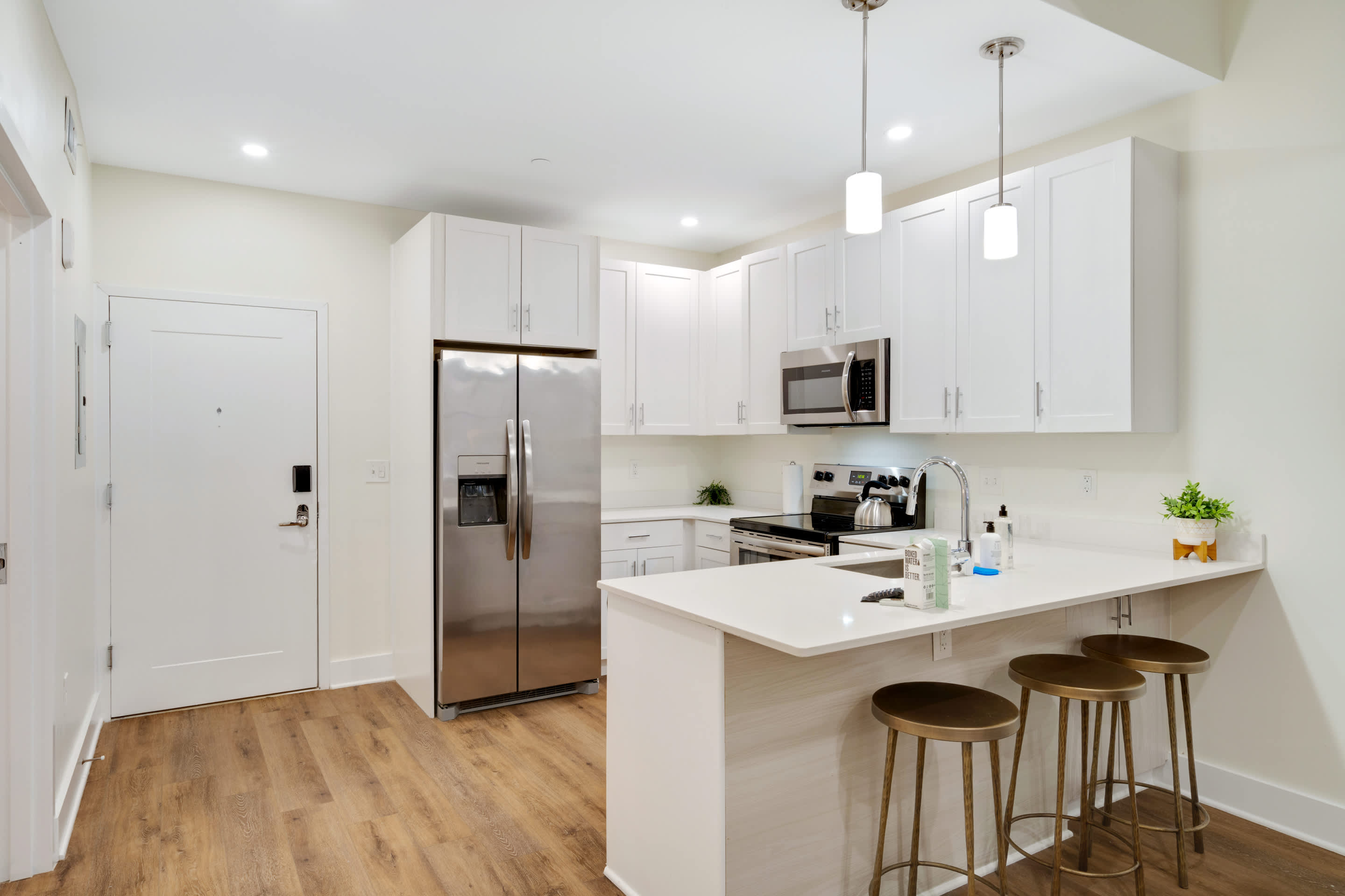 Bright, modern kitchen with white shaker cabinets, stainless steel appliances including a refrigerator, microwave, and range, and a white quartz countertop with a breakfast bar. Three bronze bar stools are positioned at the bar, illuminated by pendant lights.