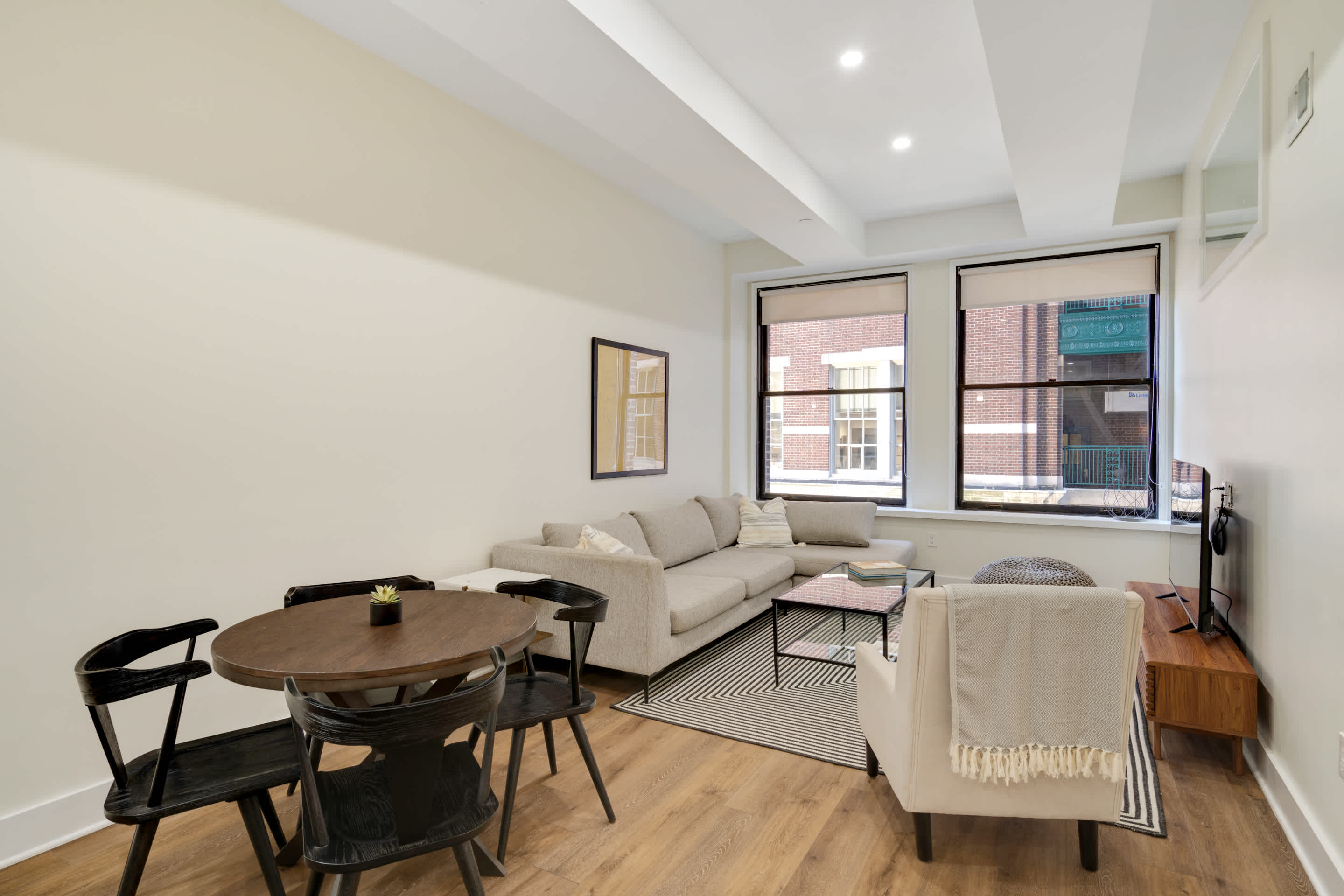 Open concept living and dining area featuring a light gray sectional sofa, a round wooden dining table with black chairs, and a black and white striped rug. Large windows provide natural light and a view of an adjacent brick building.