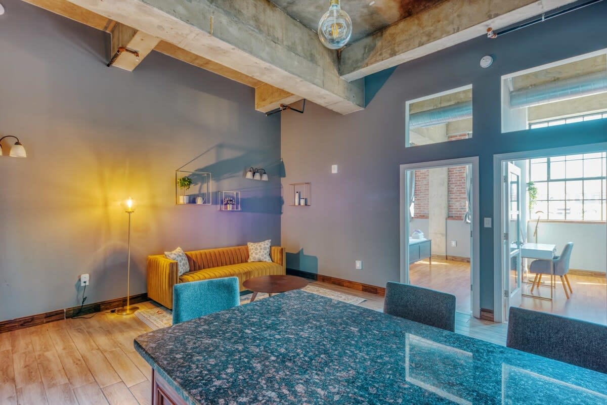 An expansive loft living area with a mustard yellow sofa, a round coffee table, and a large kitchen island with dark granite countertops and bar seating. The space features high ceilings with exposed concrete beams and industrial lighting, and light grey walls.