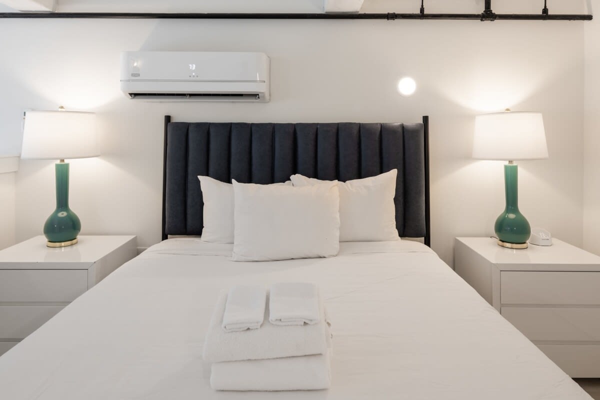 A cozy bedroom featuring a queen-sized bed with a dark gray upholstered headboard, white bedding, and two white nightstands with tall green lamps, all beneath a wall-mounted AC unit.