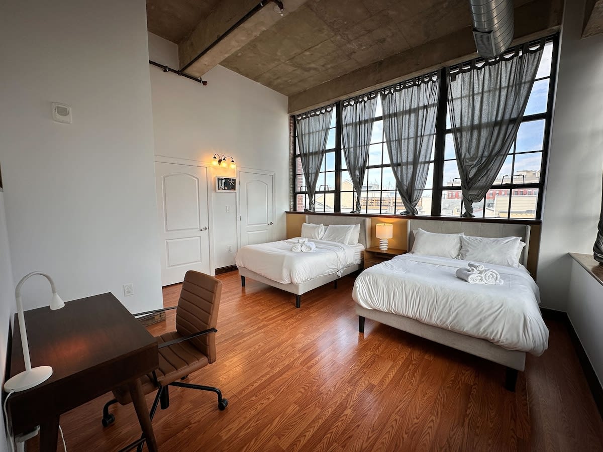 Large bedroom featuring two queen-sized beds with white linens, a brown leather office chair, and a dark wooden desk, set against a backdrop of expansive industrial-style windows with dark gray curtains.