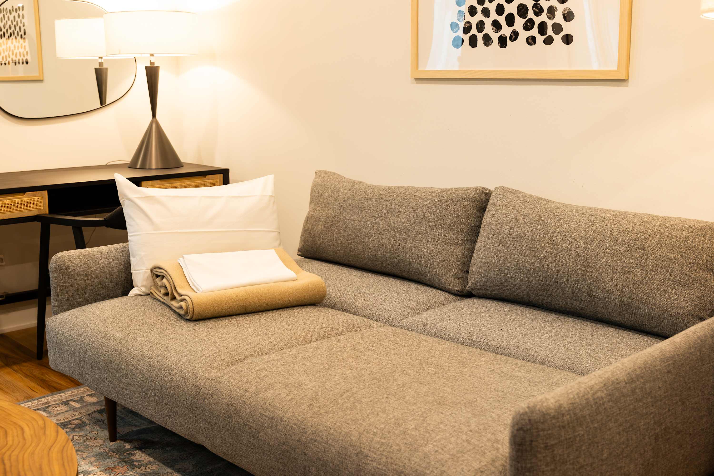 A close-up of a gray sofa with a white pillow and a beige throw blanket, positioned against a light-colored wall with framed artwork and a mirror above a dark console table with a lamp.