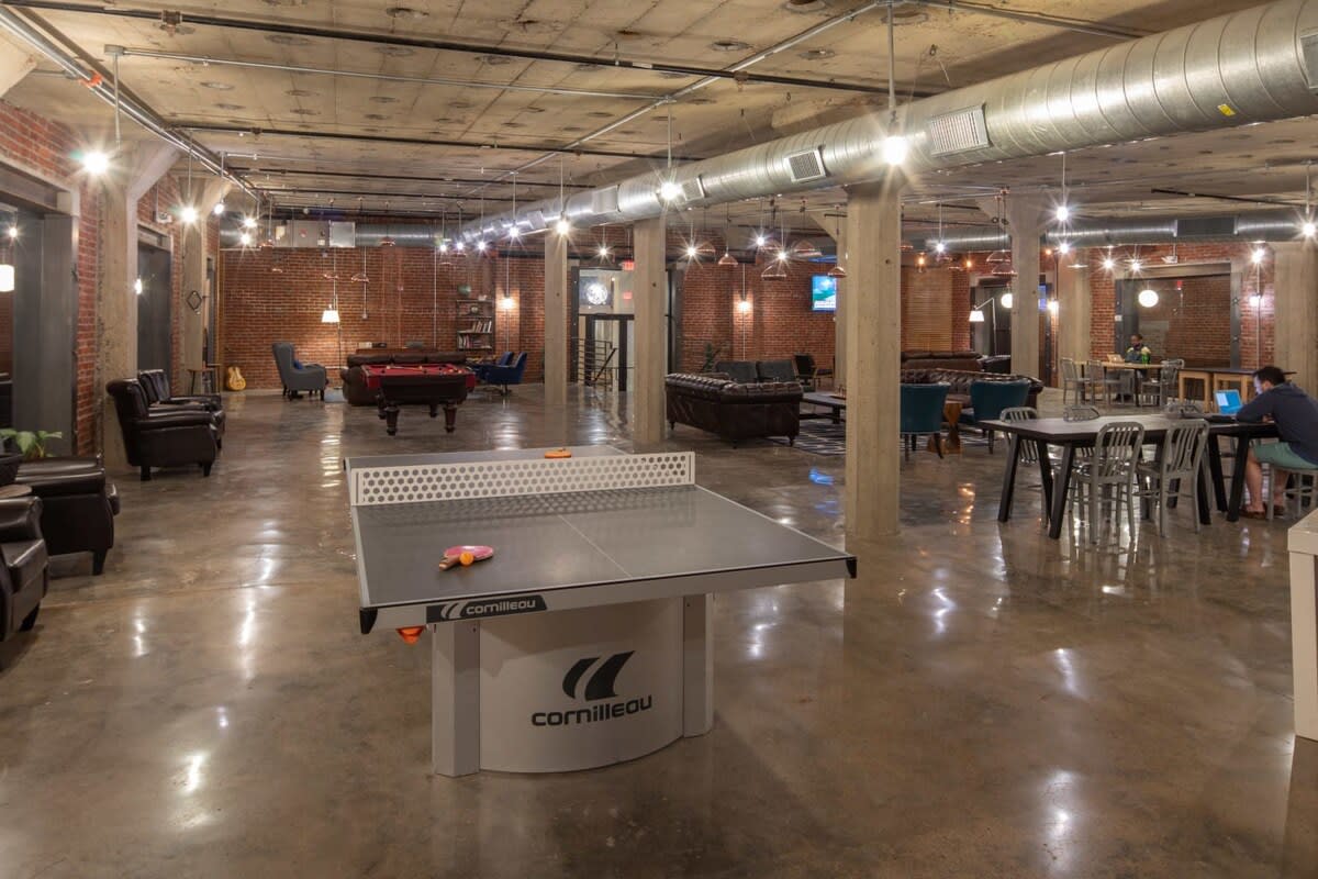 A large, industrial-chic common lounge area featuring a ping-pong table in the foreground, with various seating arrangements including leather sofas and armchairs, and a pool table in the background. Exposed brick walls, concrete floors, and visible ductwork contribute to the urban loft aesthetic.