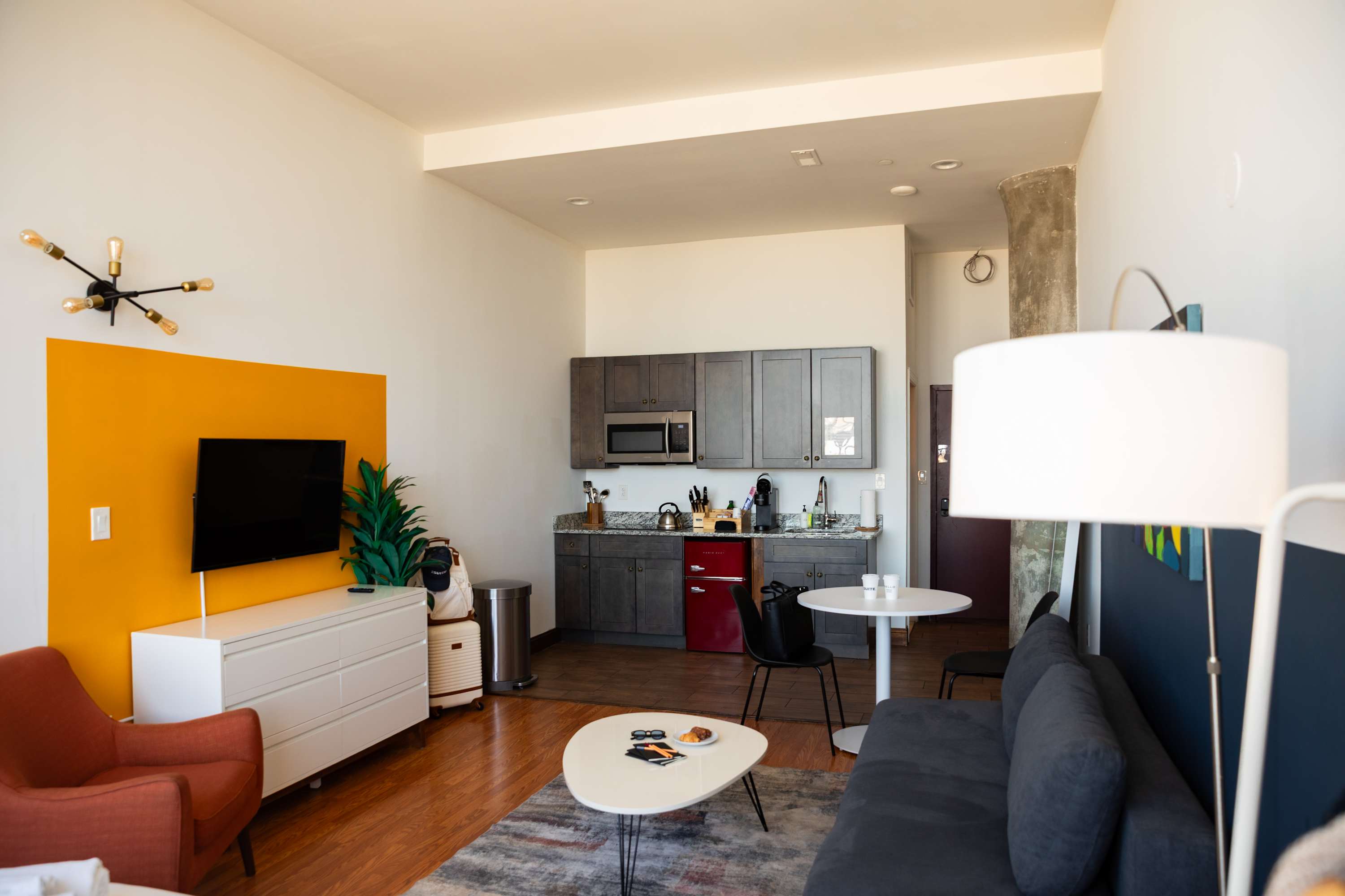 Open-concept studio apartment showcasing a compact kitchen with dark cabinets, a gray sofa, a white coffee table, and a TV mounted on an orange accent wall. The space features hardwood floors and a concrete pillar.