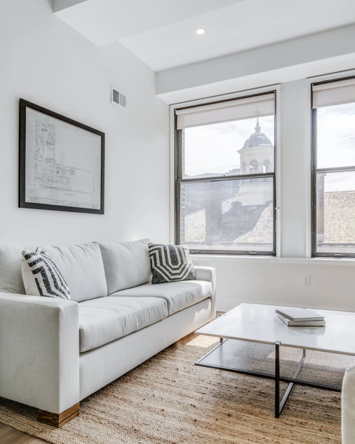 Close-up view of a light gray sofa adorned with a geometric patterned pillow, positioned next to a large window offering a glimpse of a distant architectural dome. A textured jute rug covers the hardwood floor.