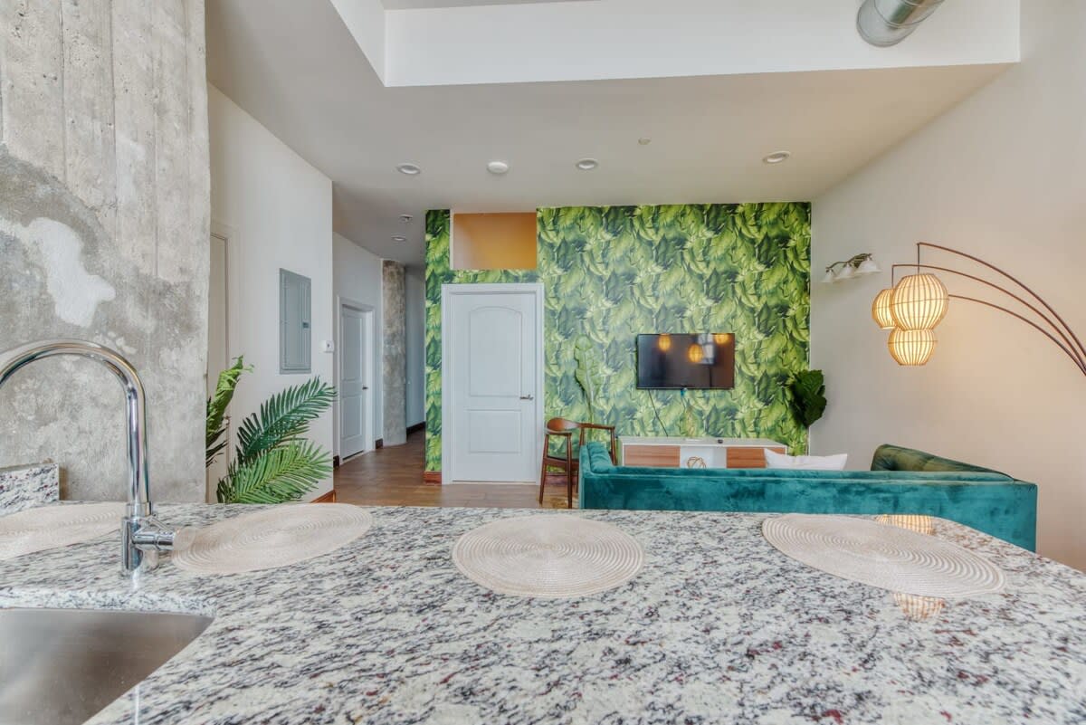An open-concept living and kitchen area, showcasing a granite kitchen island with a sink in the foreground, leading to a living space with green banana leaf wallpaper and a teal sofa.