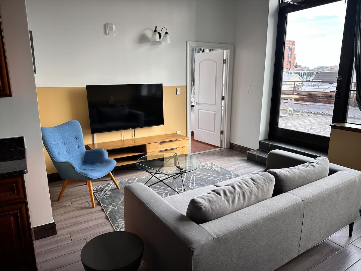 Bright living room with a light gray sofa, a blue accent chair, and a large flat-screen TV on a wooden console, set against a white and mustard yellow wall, with a glass coffee table on a patterned rug.