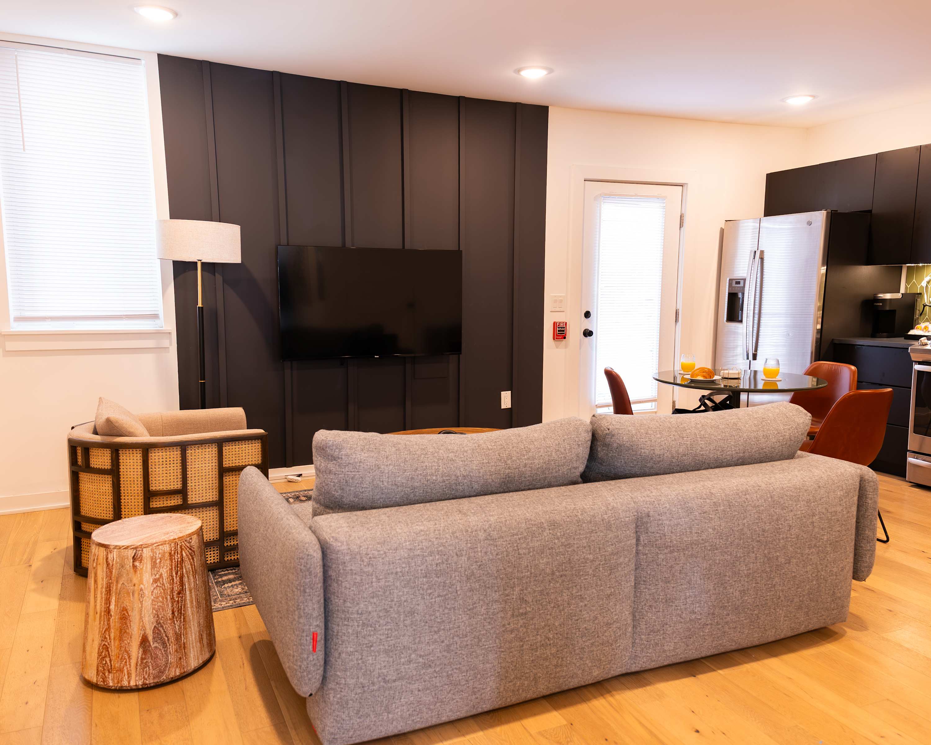 A modern living room with light wood floors features a gray sofa facing a wall-mounted TV above a dark accent wall, with a round wooden coffee table and a dining area with a glass-top table and brown chairs visible in the background.
