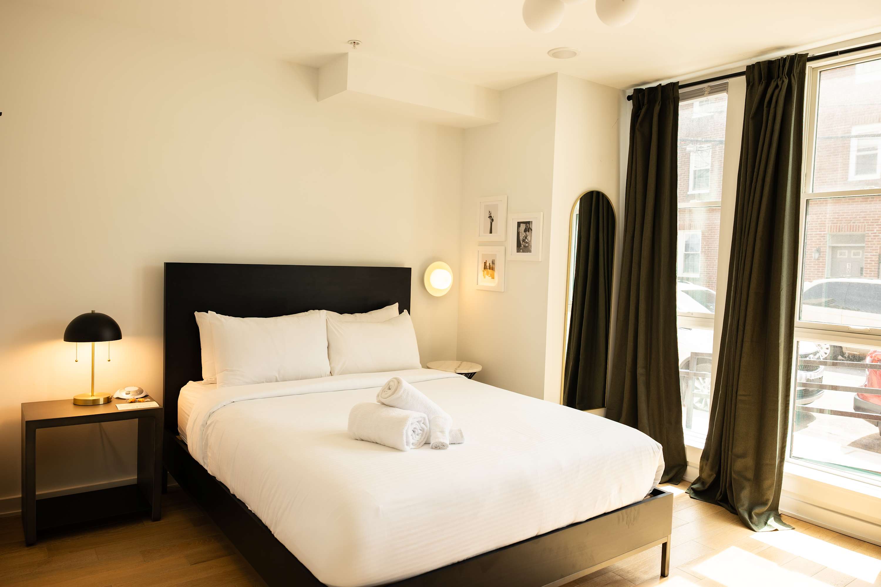 A bright bedroom features a queen-sized bed with white linens and a black headboard, flanked by a black nightstand with a lamp, and large windows with dark green curtains offering natural light.