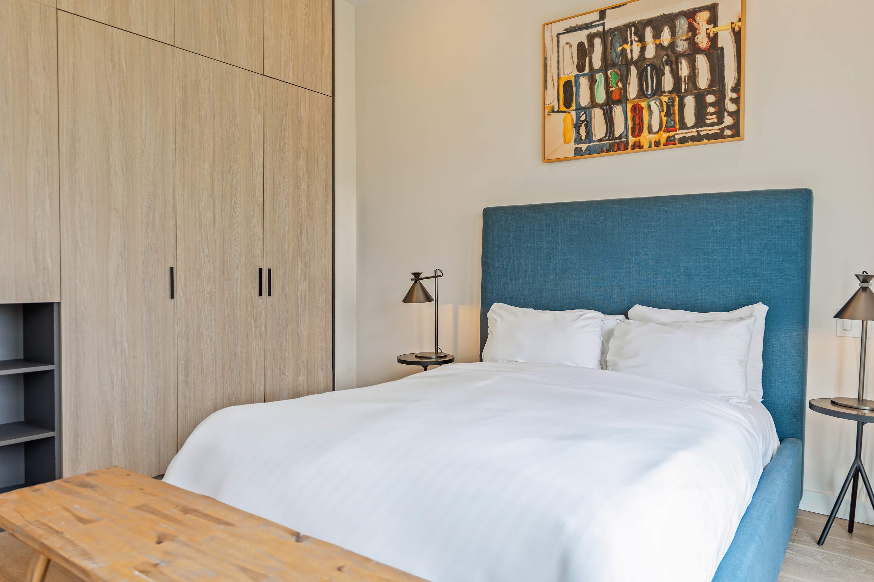 Contemporary bedroom with a blue upholstered queen bed, white linens, and black metal nightstands with lamps. A large light wood built-in wardrobe with black handles and open shelving is on the left, and abstract artwork hangs above the bed.