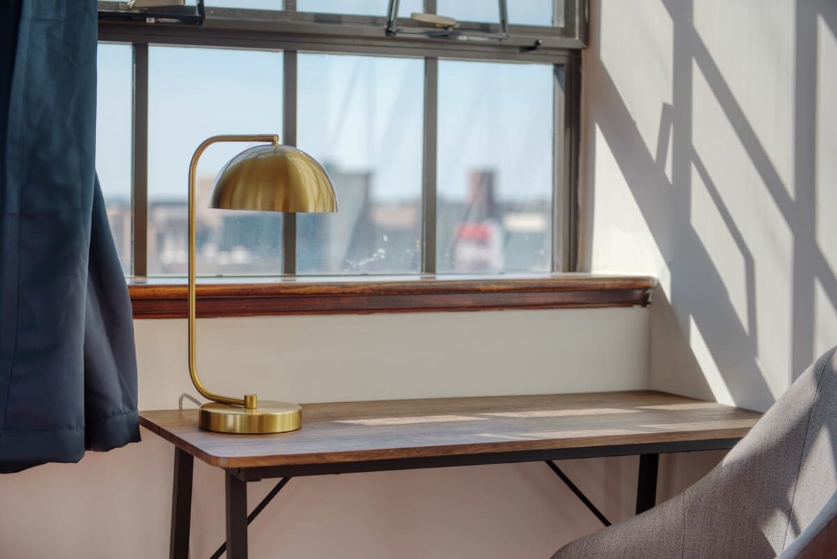 Dedicated workspace with a wooden desk and a modern brass lamp, positioned by a large industrial window offering natural light and a partial city view.