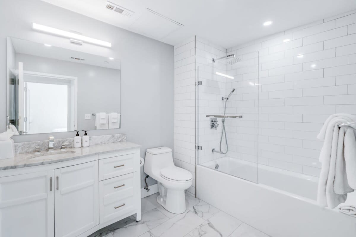 Clean white bathroom featuring a single vanity with a marble countertop and white cabinet, a toilet, and a bathtub with a glass shower screen. White subway tiles line the shower walls, and marble-look floor tiles complete the space.