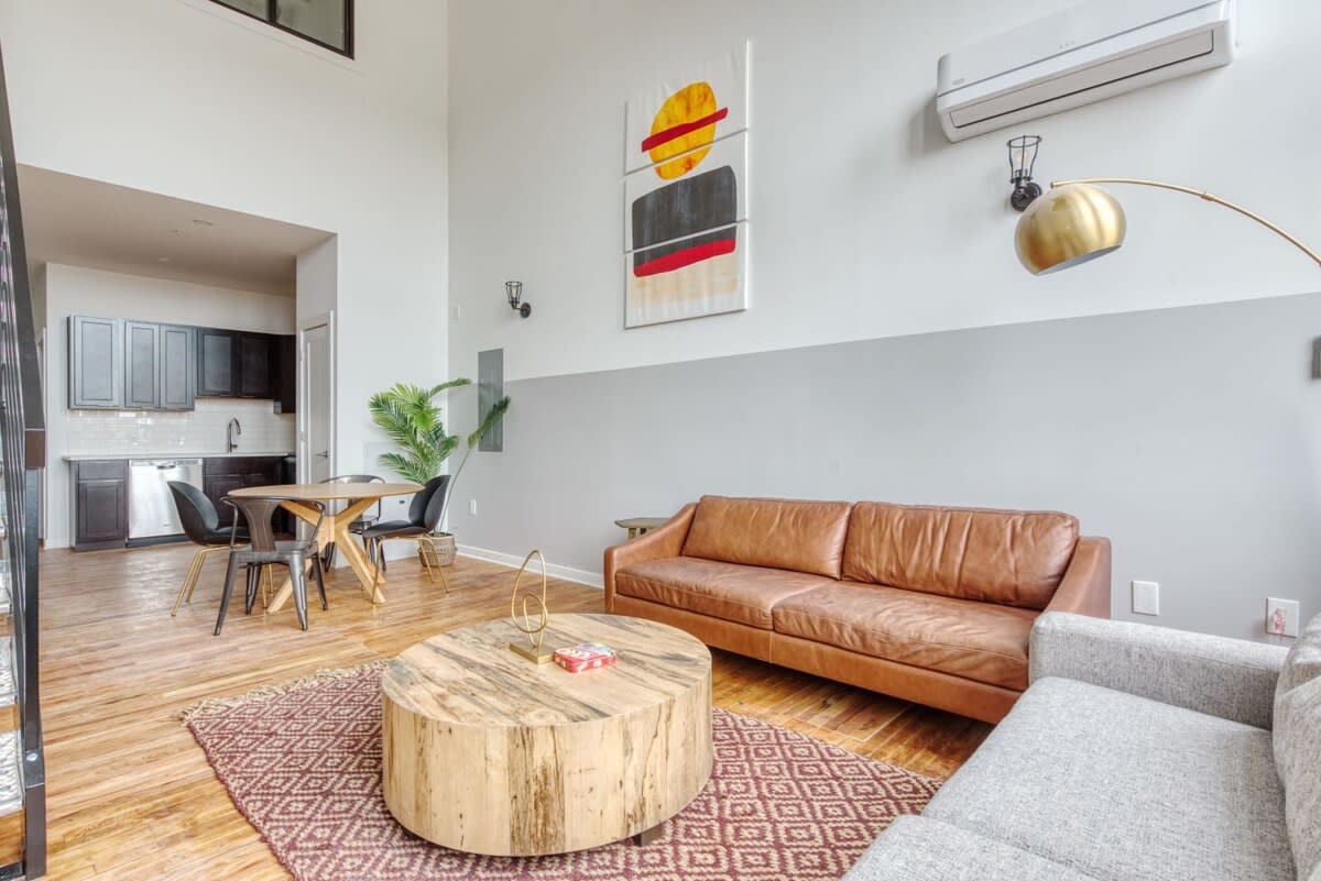 Spacious open-plan living area with a brown leather sofa, a grey sofa, and a round wooden coffee table on a patterned rug, adjacent to a dining area and a modern kitchen with dark cabinetry.