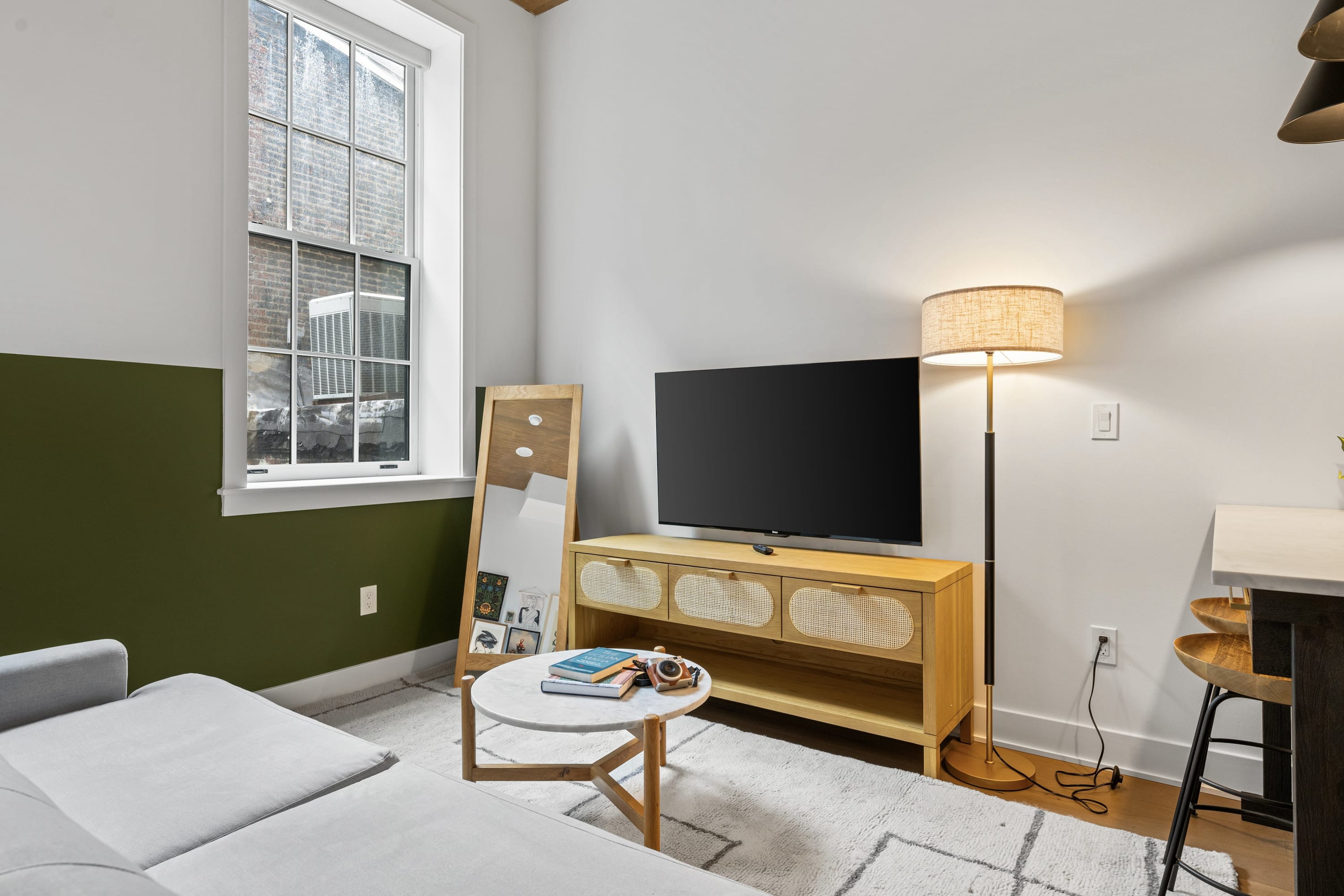 A detailed view of the living area showcasing a large flat-screen TV on a light wood console with woven cabinet fronts. A full-length mirror leans against the wall, reflecting the room, next to a window with an air conditioning unit.