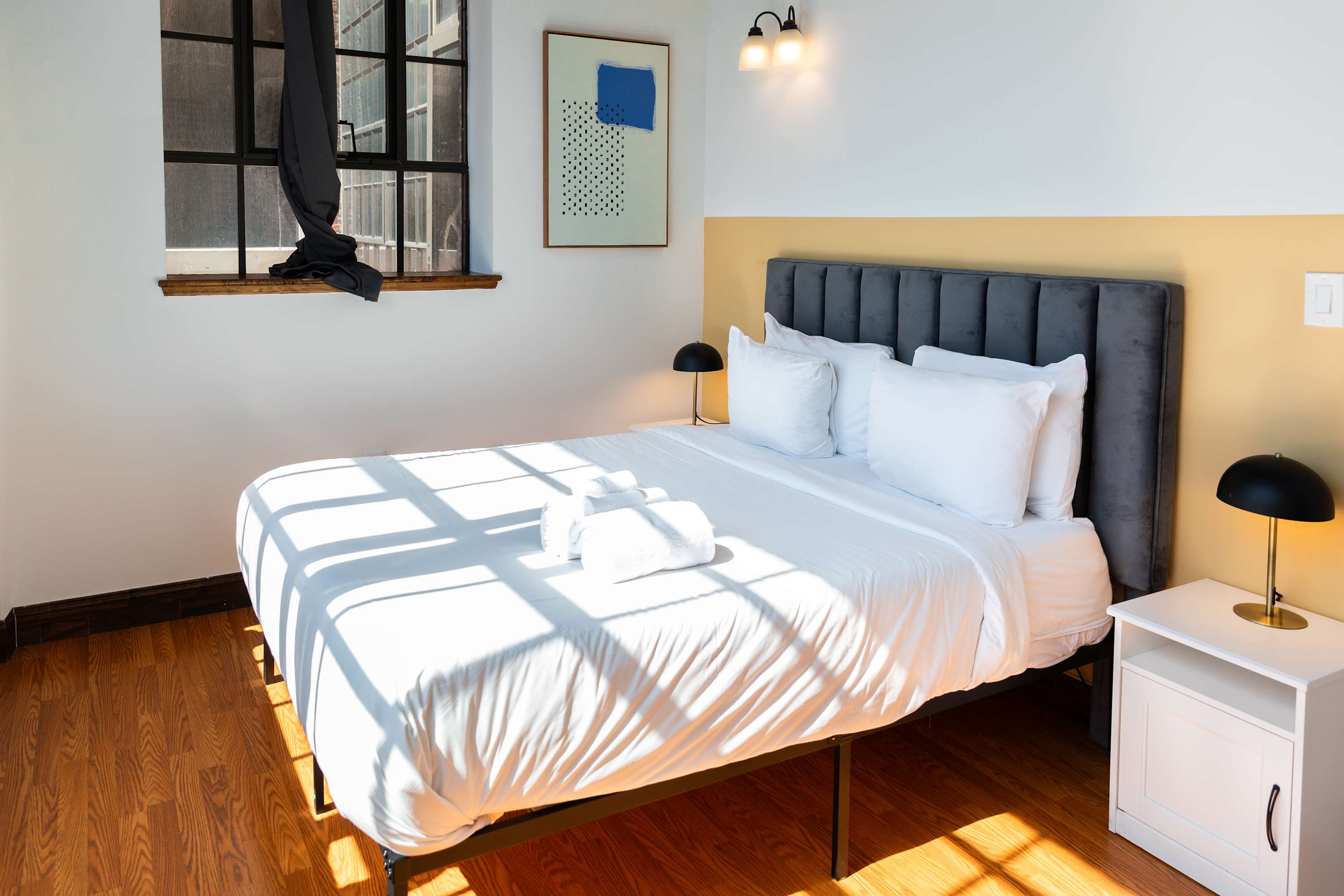 Bedroom with a queen-sized bed featuring a grey upholstered headboard, white bedding, and two black table lamps on white nightstands, set against a two-tone wall (white and mustard yellow) with an industrial-style window.
