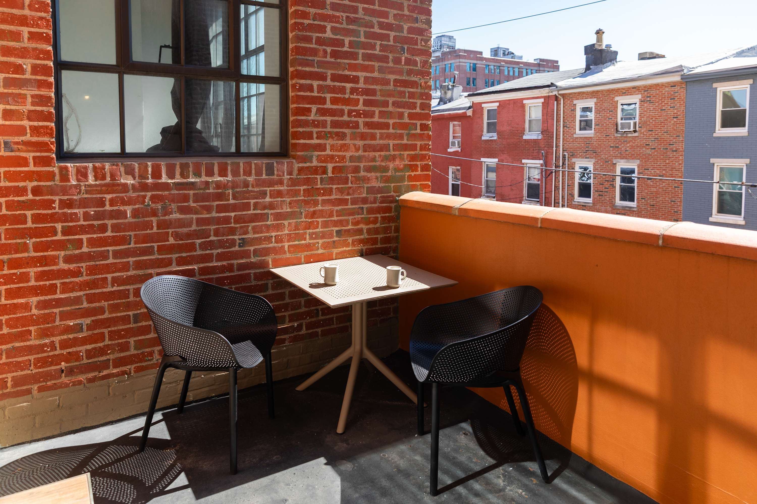 Outdoor balcony with exposed brick walls, featuring a small square table and two modern black chairs, with two mugs on the table, offering a view of adjacent buildings.