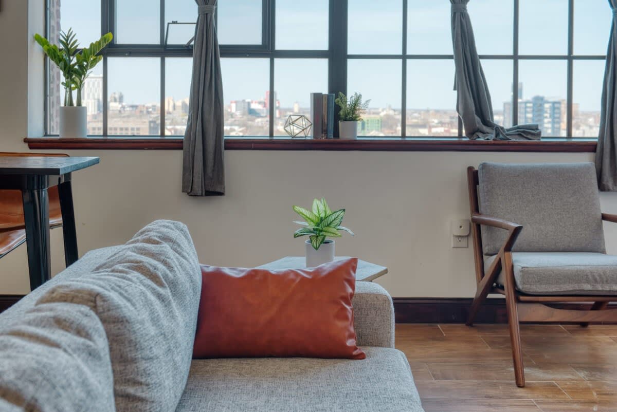 Close-up of the living room showing a light gray sofa with a brown throw pillow, a side table with a plant, and a mid-century modern armchair, all bathed in natural light from large windows.