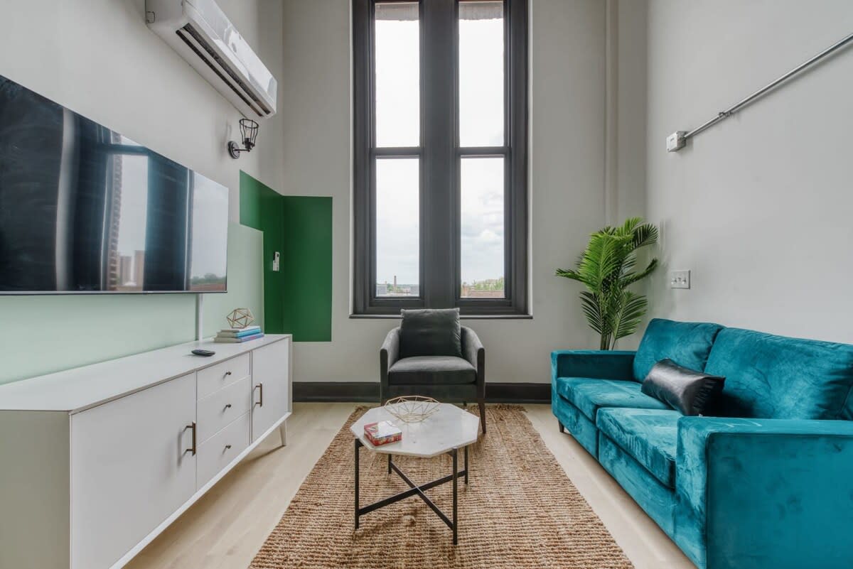 Modern living room with a teal velvet sofa, a gray armchair, and a white media console with a large TV. A jute rug covers light wood floors, and tall dark-framed windows provide natural light. A green accent wall is visible behind the TV.