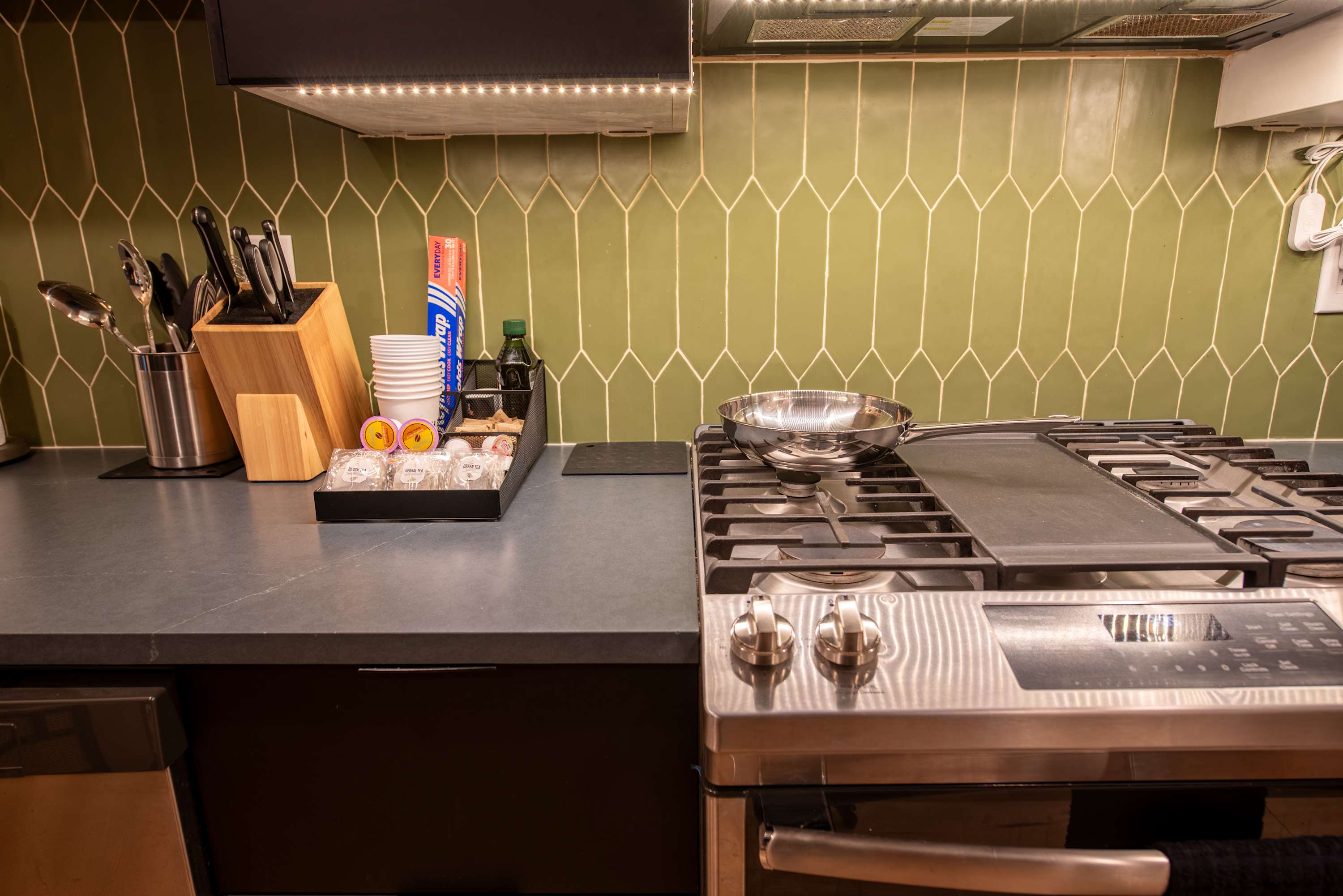 A close-up of a modern kitchen counter with a gas range, stainless steel appliances, and a distinctive green picket tile backsplash, featuring a coffee station and cooking utensils.