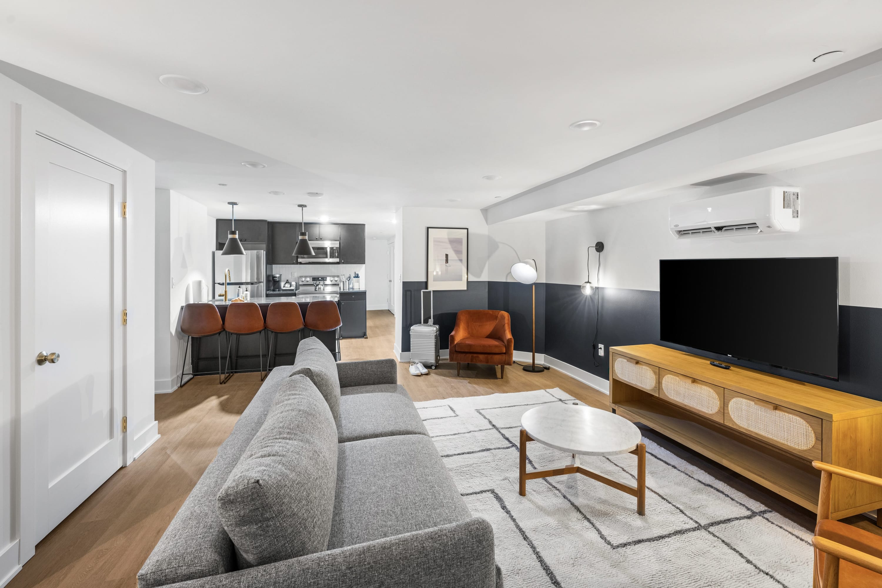 A wide shot of the modern living room, showing a gray sofa, a patterned rug, and a wooden coffee table. In the background, the kitchen with dark cabinetry and bar stools is visible, connecting the open-concept space.