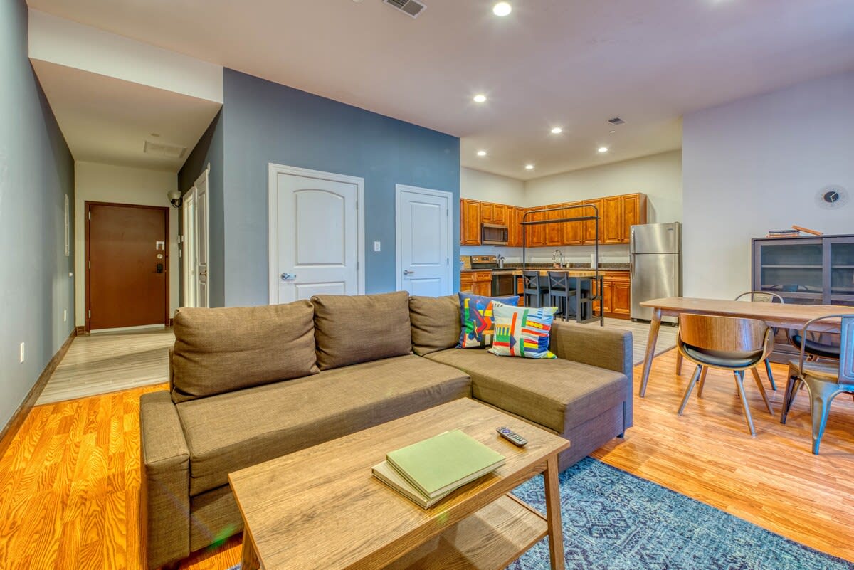 Open-concept living space showcasing a brown sectional sofa, a wooden coffee table, and a dining area with a wooden table and chairs. The kitchen is visible in the background.