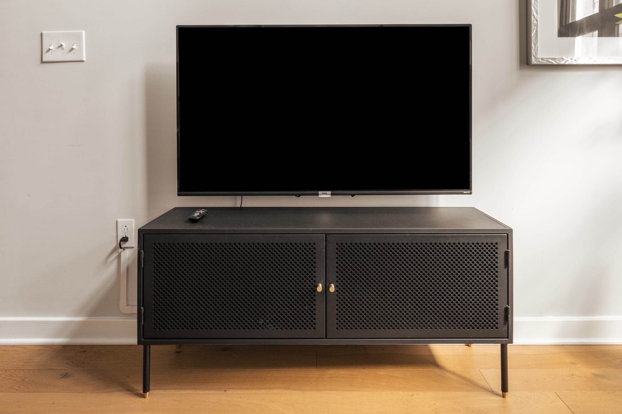 A close-up of the TV area in the living room, showing a wall-mounted flat-screen television above a black perforated metal media console with two doors. The console has gold handles and thin legs.