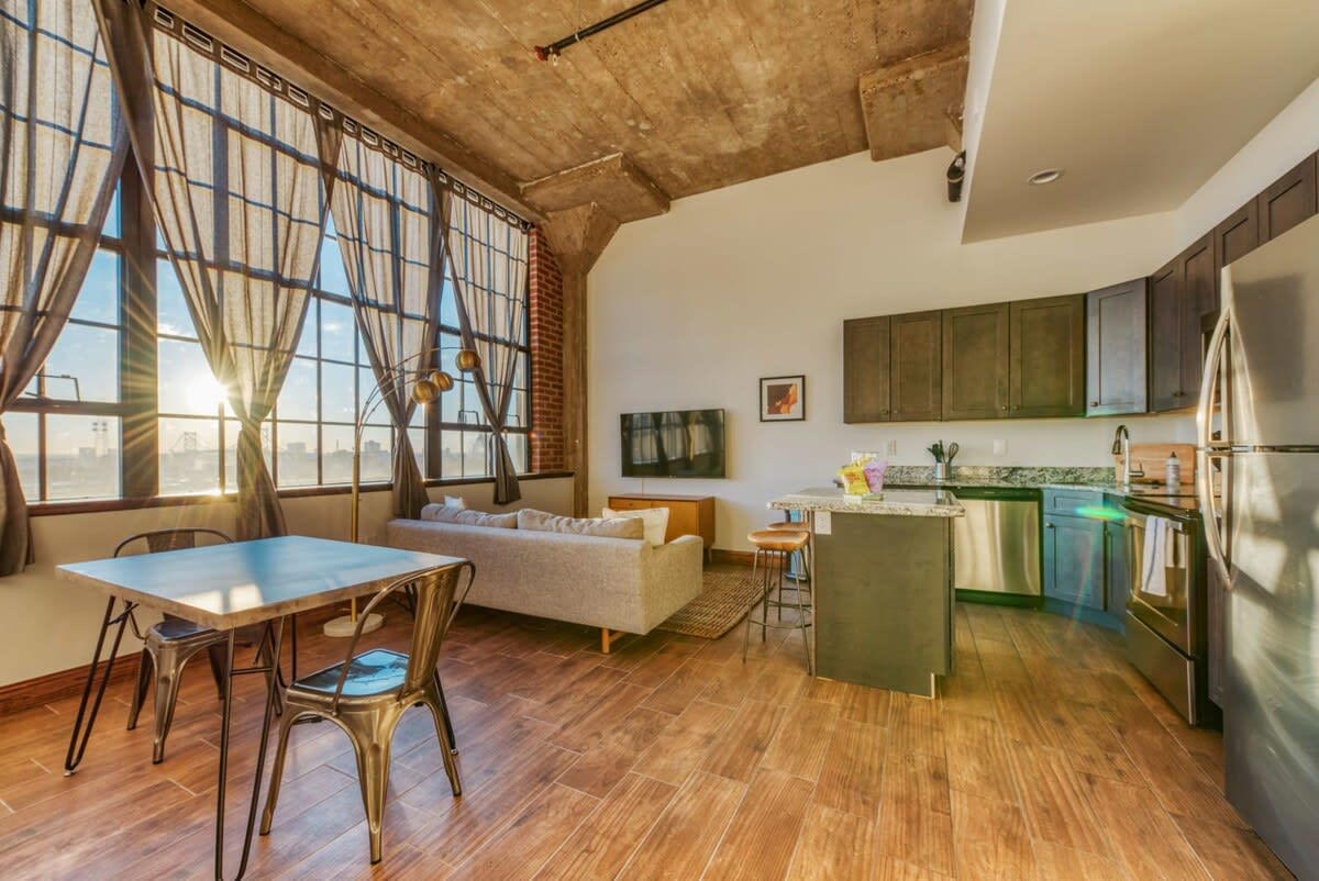 An expansive open-concept living, dining, and kitchen area with high ceilings, exposed concrete, and large industrial windows offering panoramic city views. The space features hardwood floors, a light gray sofa, and a dining table with metal chairs.