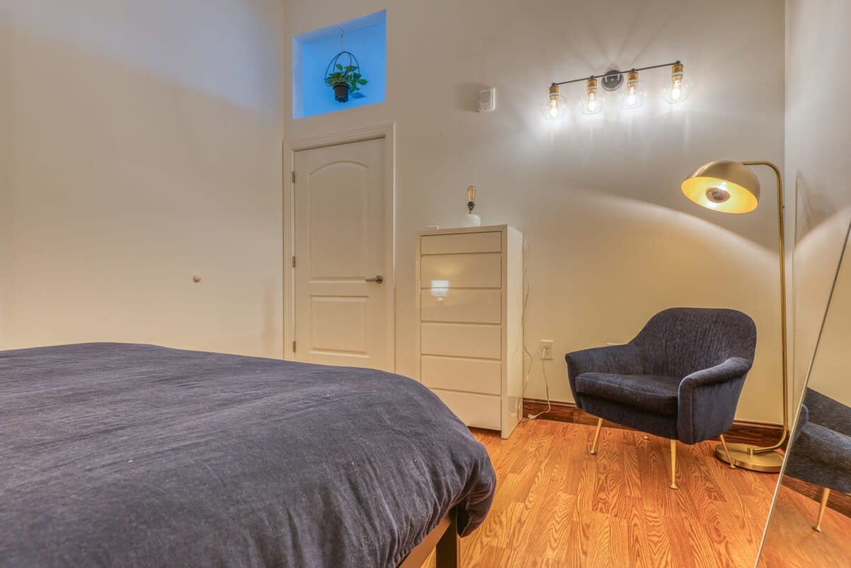 Another view of the bedroom shows the dark blue bed, a white dresser, and a dark blue armchair with gold legs. A tall gold floor lamp and a full-length mirror are also present, with light wood flooring throughout.