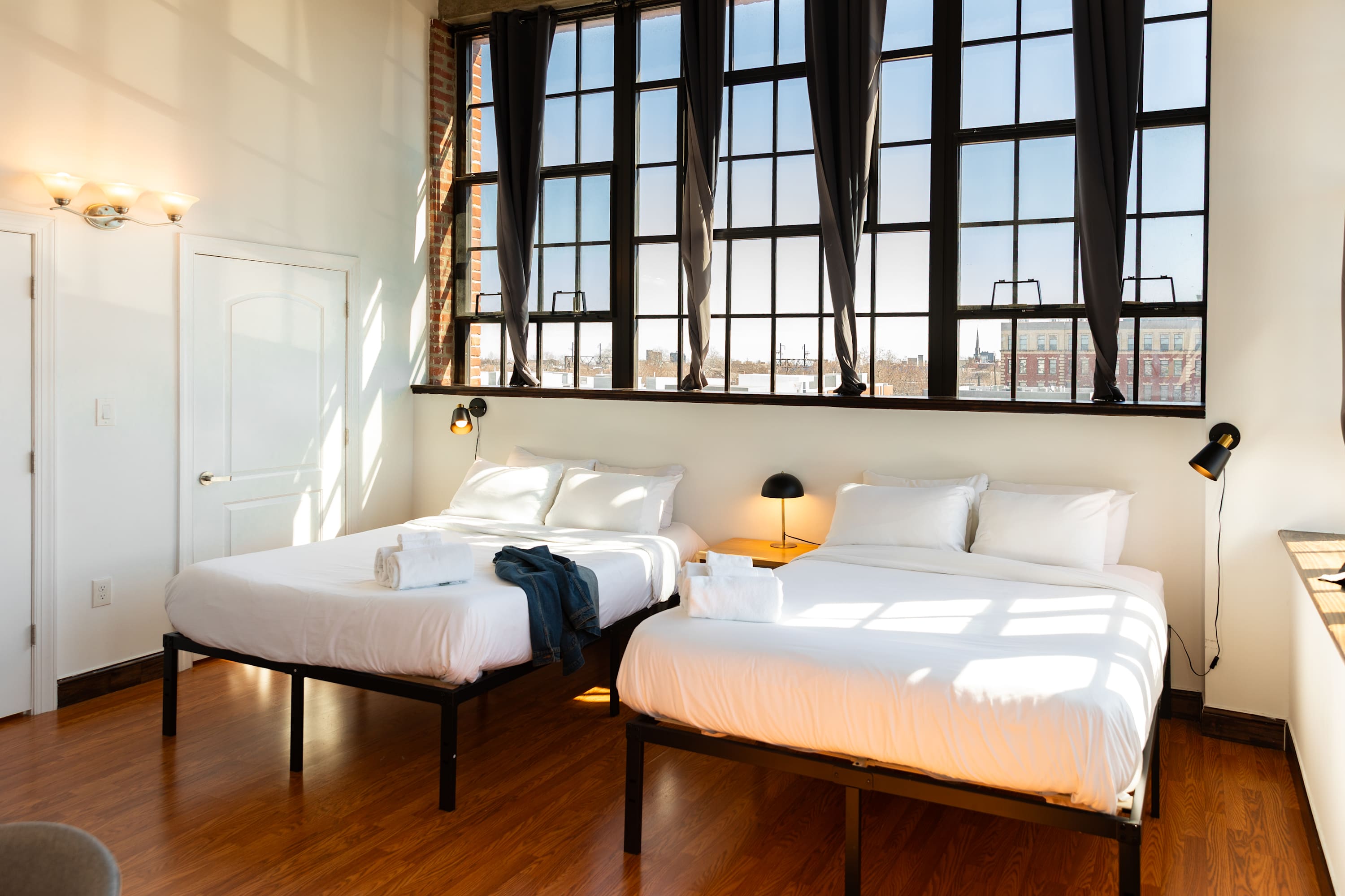 A spacious bedroom with two queen-sized beds, hardwood floors, and a wall of large industrial windows with dark curtains. Each bed has a wall-mounted reading lamp and white bedding.
