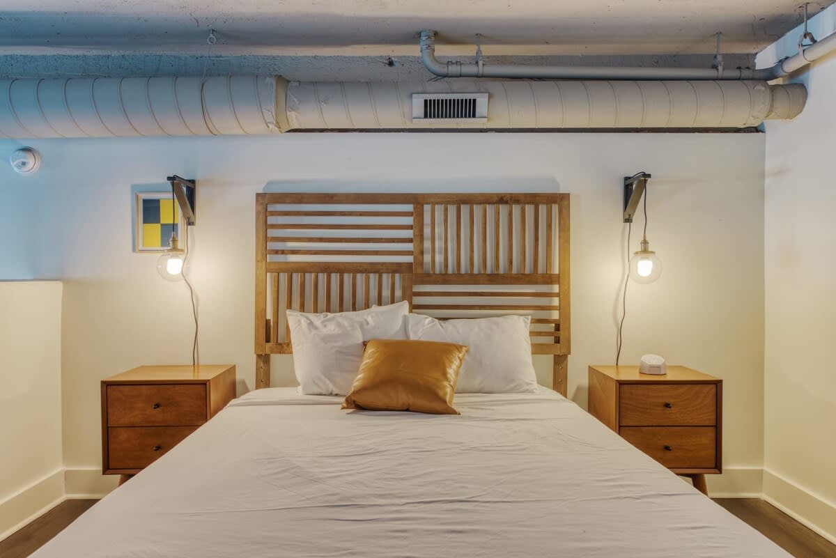 Cozy bedroom with a wooden slatted headboard, two matching wooden nightstands with industrial-style wall sconces, and a bed with white linens and a mustard yellow accent pillow. Exposed ductwork is visible on the ceiling.