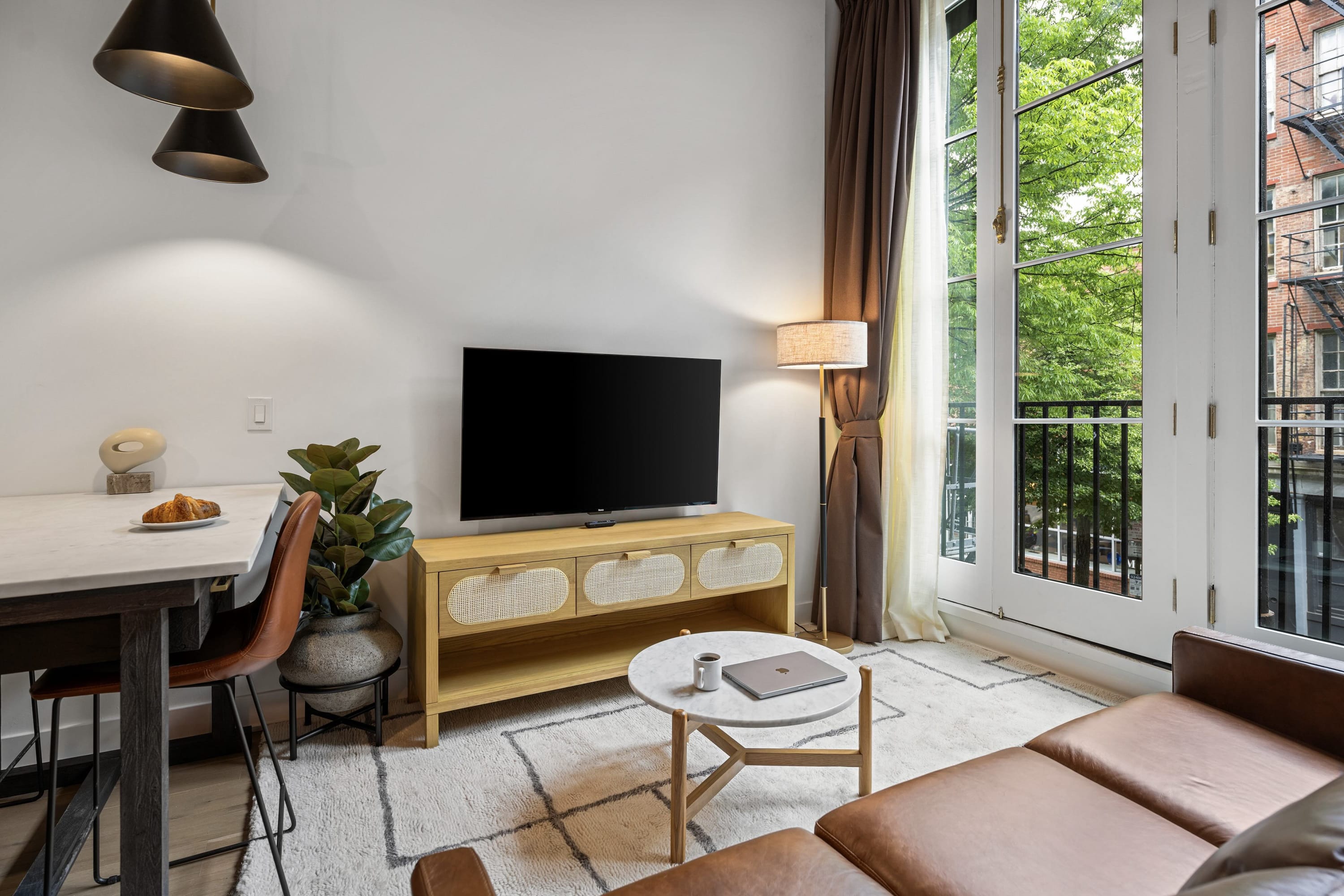 A bright living area featuring a brown leather sofa, a large flat-screen TV on a light wood console, and a round marble-top coffee table. French doors with a Juliet balcony offer a view of the city outside.