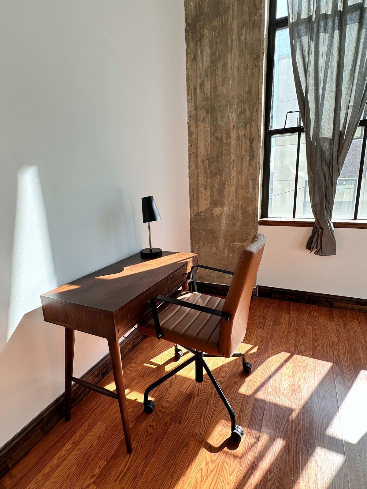 Dedicated workspace featuring a dark wood desk with a black and brown office chair, illuminated by natural light from a large window. A small black desk lamp is also present.