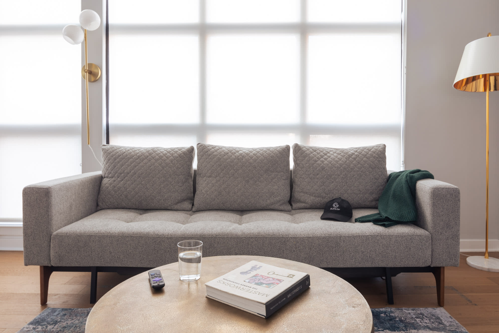 Close-up of a light gray sofa with quilted cushions, a round brass coffee table with a book and glass of water, and a dark green throw blanket. Large windows with white roller blinds provide bright natural light.