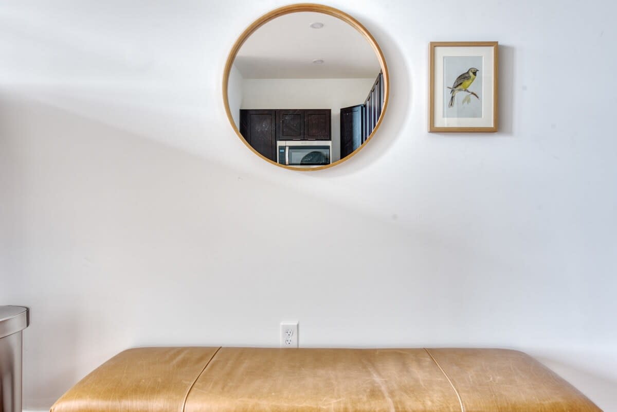 Minimalist entryway featuring a round wooden-framed mirror reflecting the kitchen, a framed bird print, and a light brown leather bench against a white wall.