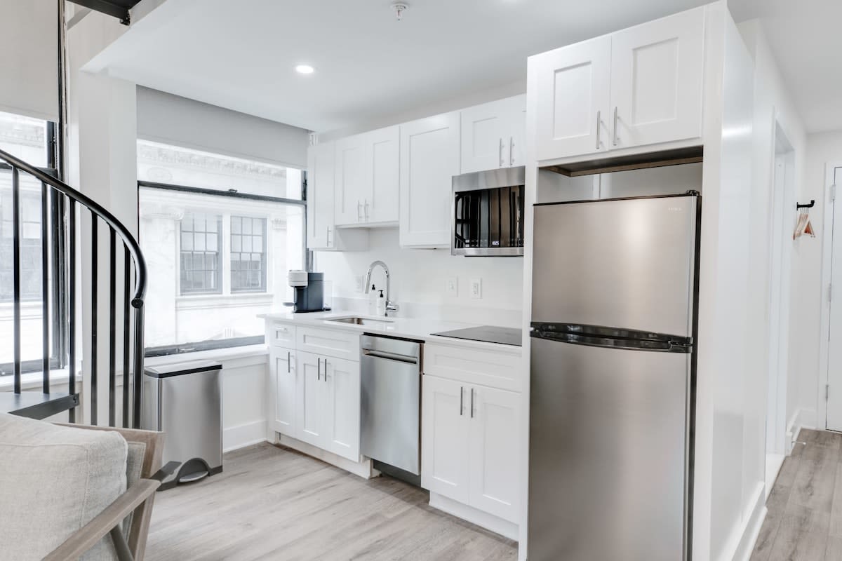 A compact, modern kitchen with white shaker cabinets, stainless steel appliances including a refrigerator, microwave, and dishwasher, and a white countertop. A window is visible next to a stainless steel trash can.